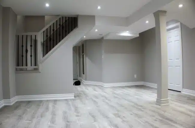 A modern finished basement featuring grey laminate flooring, white trim, recessed lighting, and a staircase adorned with black iron railings.