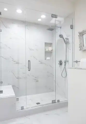 A modern, spacious bathroom featuring a glass-enclosed shower with a marble interior, a mounted showerhead, built-in shelving, and a bench. The area is well-lit with recessed lighting.