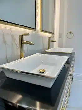 A modern bathroom features two rectangular white sinks with gold faucets on a black countertop, and a large mirror with gold accents.